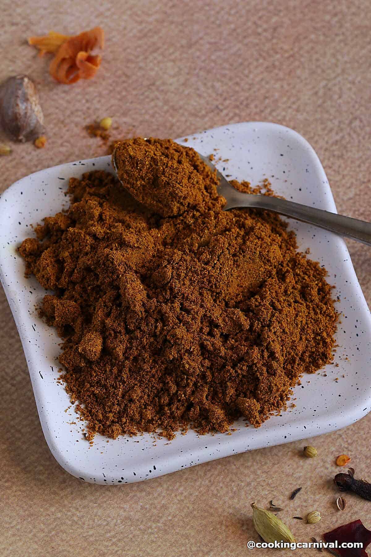 Homemade Biryani Masala powder on a white square plate with a spoon.