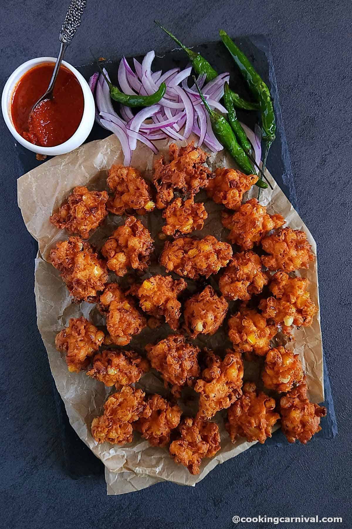 Corn pakoda on a black slate with onion, fried chilies and garlic chutney.