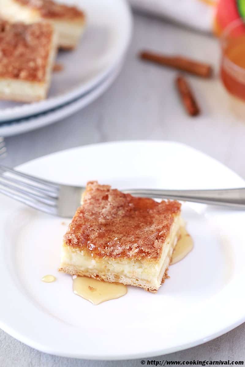 Sopapilla Cheesecake Bars with honey on white plate, fork, cinnamon sticks on the side