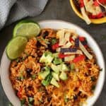 Enchilada Rice in white bowl topped with avocado, tortilla chips and lemon wedges on the sides