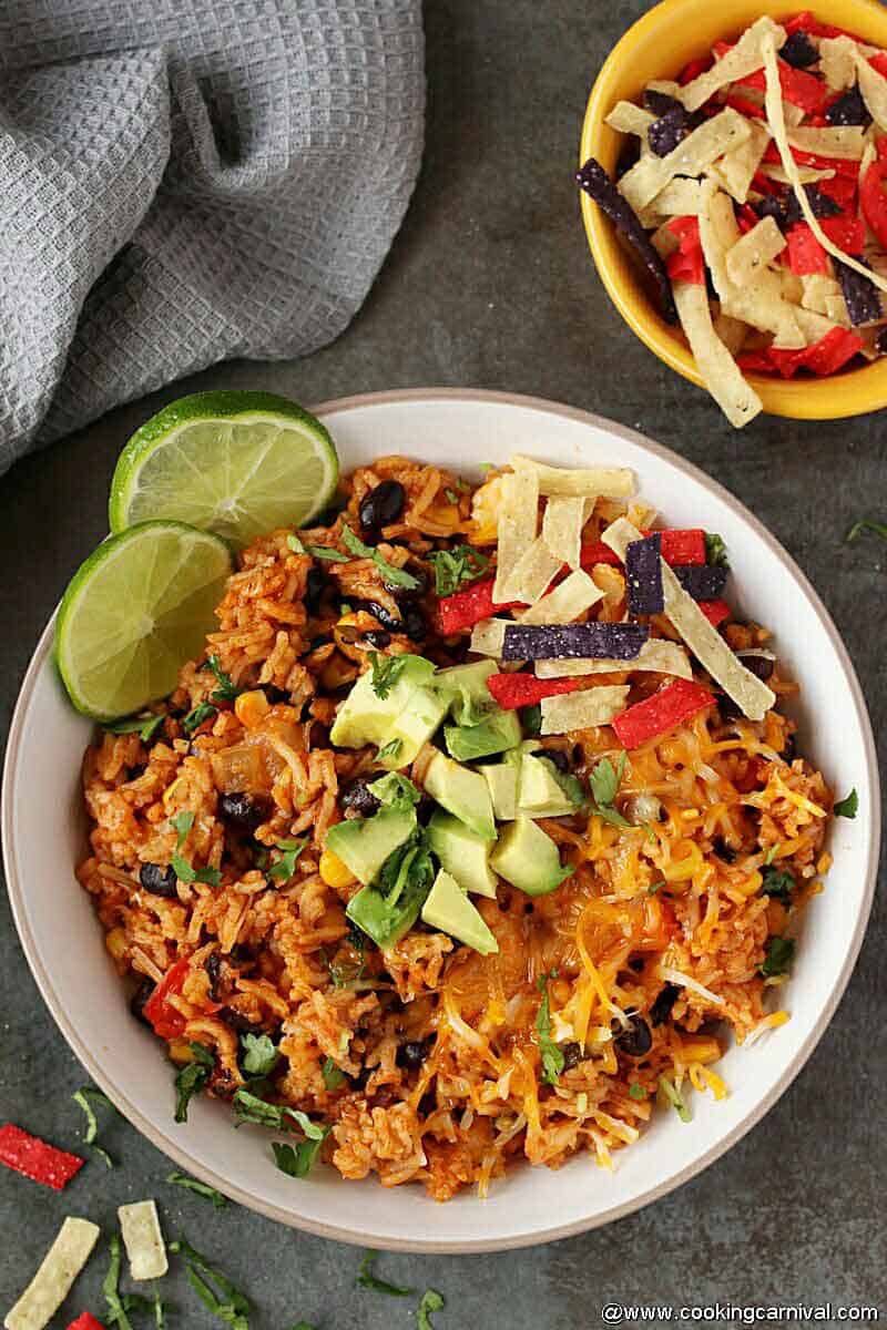 Enchilada Rice in white bowl topped with avocado, tortilla chips and lemon wedges on the sides