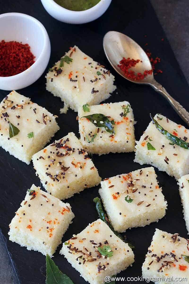 Khatta dhokla on a black cheese board, chili powder and oil on the sides