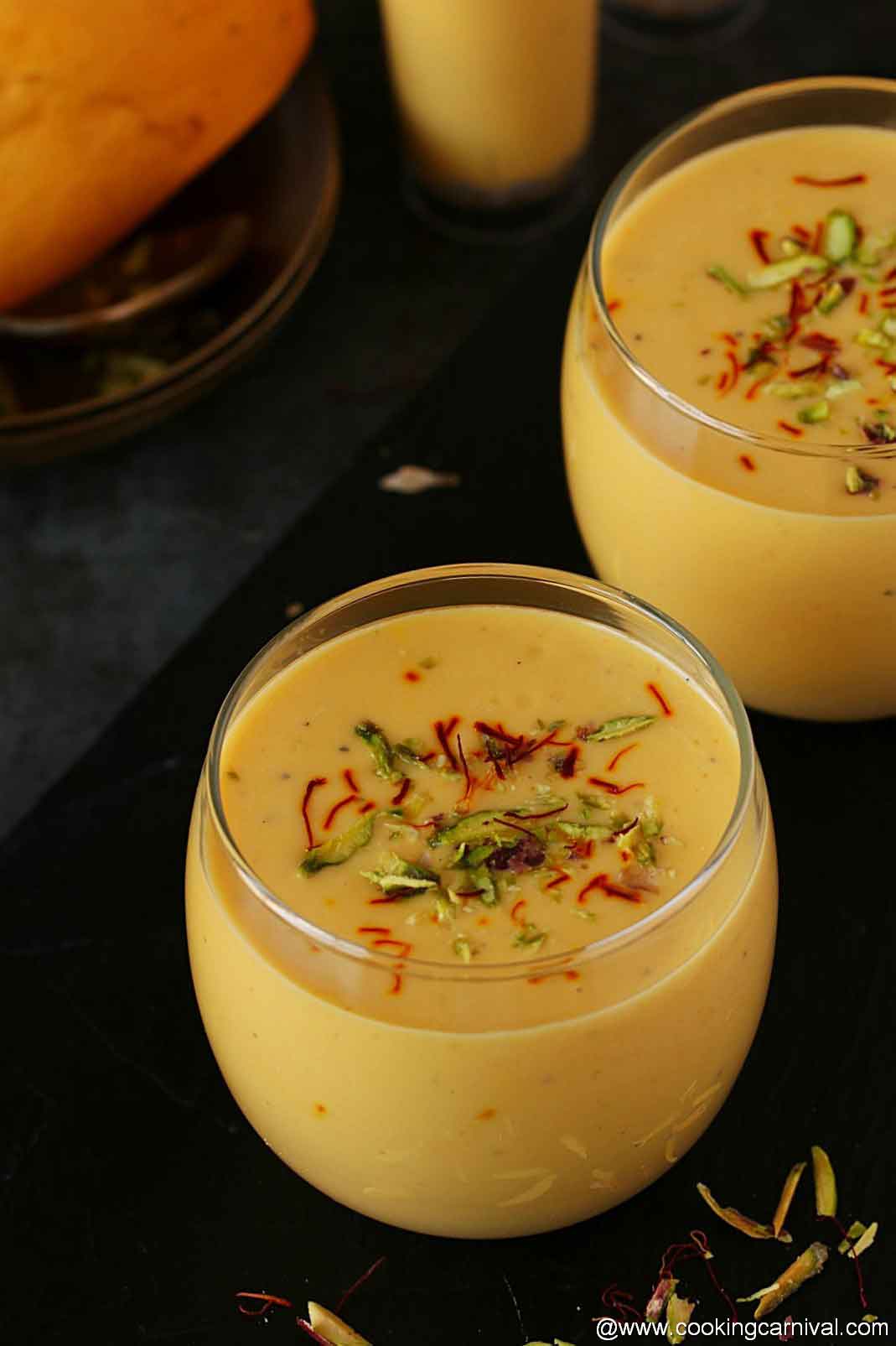 Mango lassi served in 2 glasses, garnished with saffron, pistachios and cardamom