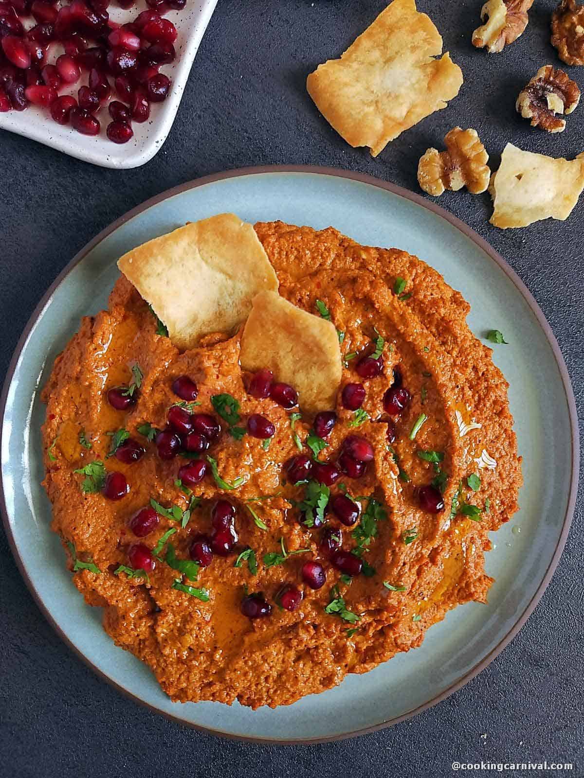 Muhammara dip on a plate garnished with pomegranate, olive oil and cilantro. Served with Pita chips.