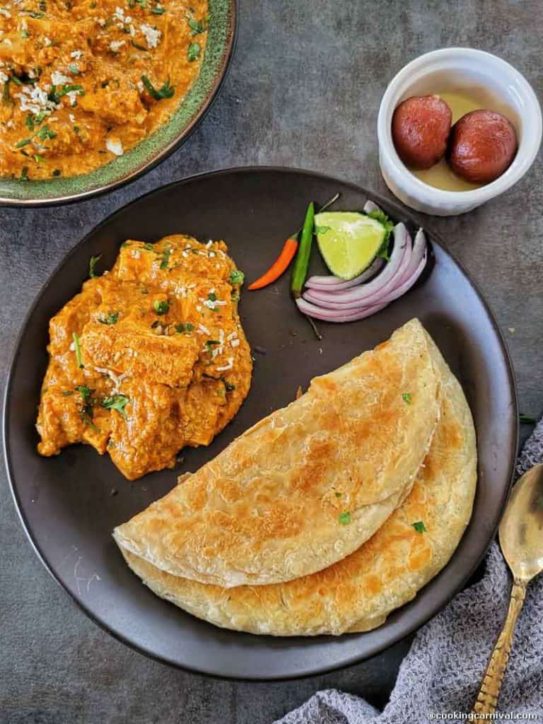 Paneer curry, paratha, onion, chii, gulab jamun and lime on a black plate