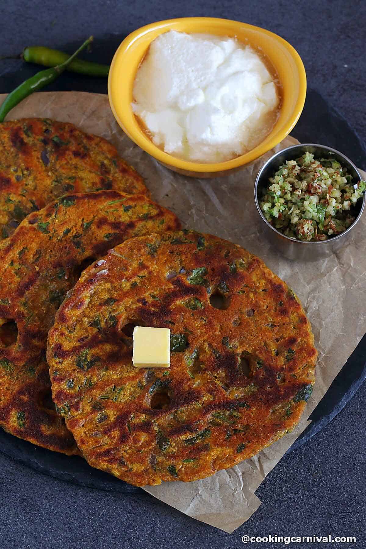 Thalipeeth served with curd, makhan, and thecha on a black slate. 