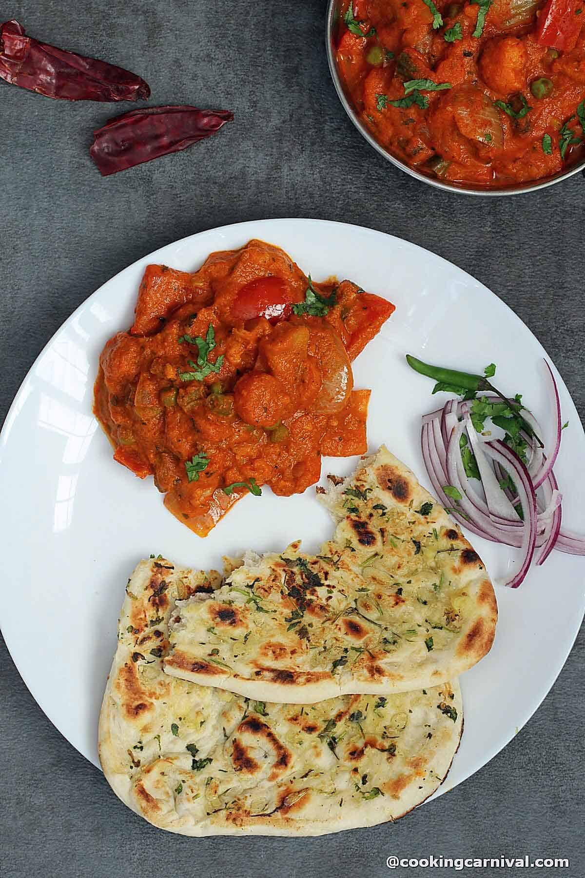 Veg Kolhapuri served with garlic naan, onions and green chili on a white plate.