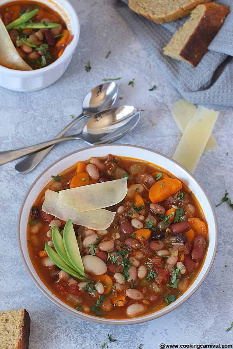 Vegetarian 15 beans soup in white bowl with Parmesan cheese, cilantro and avocado, 2 spoons and some crusty bread on the side 