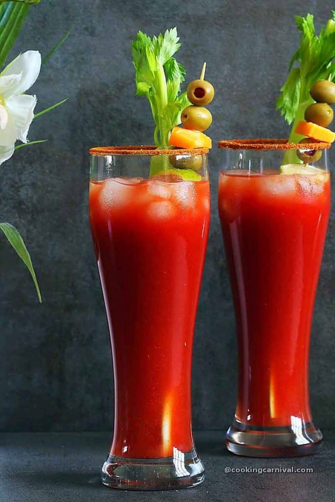 Virgin Bloody Mary drink in 2 tall glasses, garnished with celery, olives, lime and cheese