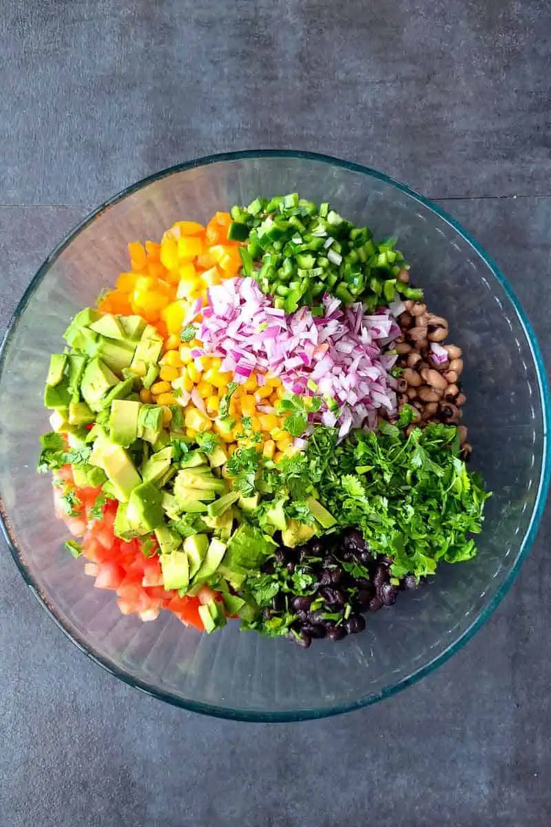 a bowl filled with black beans, black eyed peas, bell pepper, onion, corn, avocado, tomato, cilantro to make cowboy caviar salad