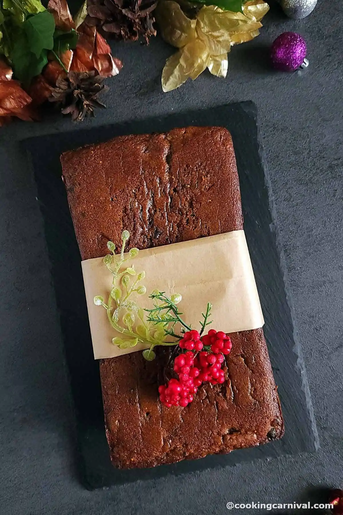 Eggless Christmas Fruit cake loaf on black board, decorated with holiday ornaments.