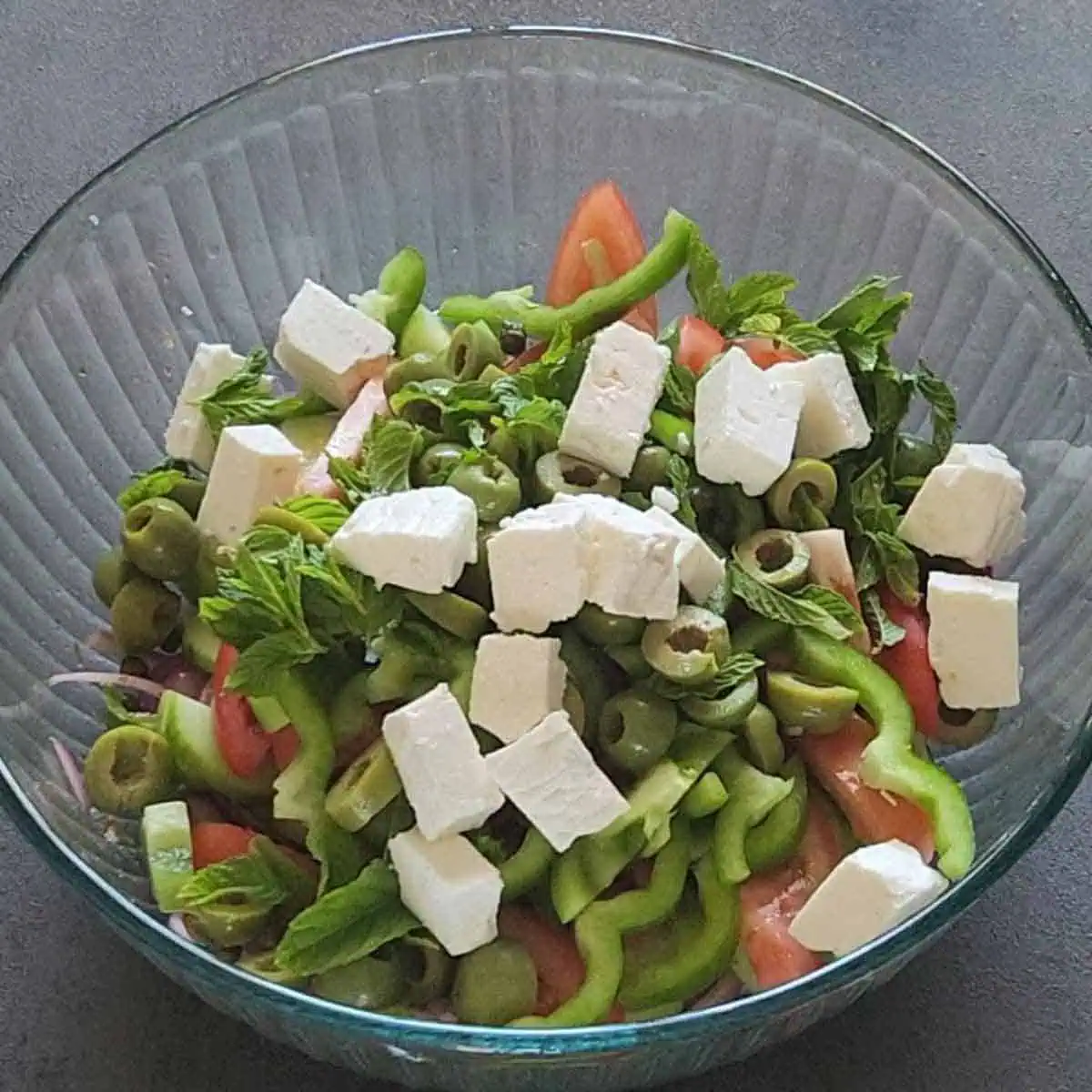 Bell pepper, cucumber, tomato, feta cheese, olive, mint and dressing in a bowl.