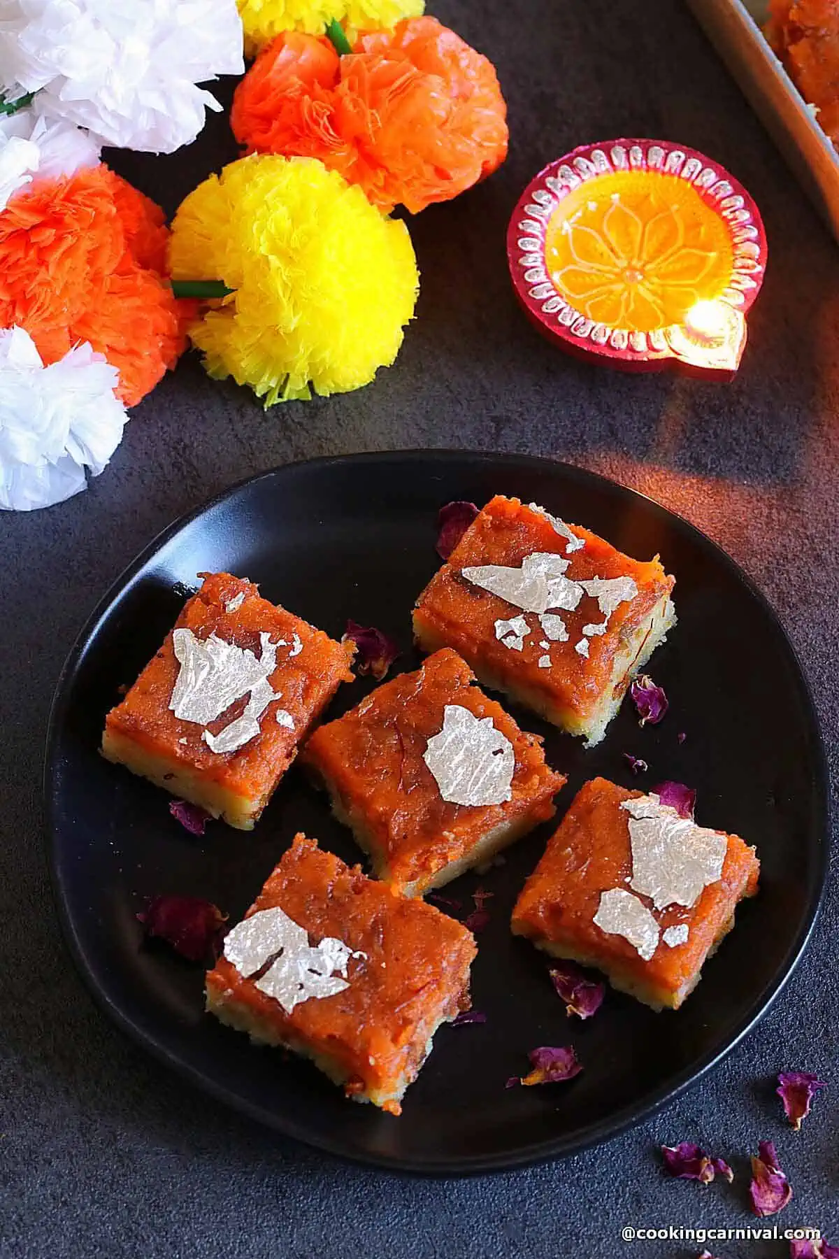 Makhana gulkand burfi on a black plate, diya and flowers on the side for decoration. 