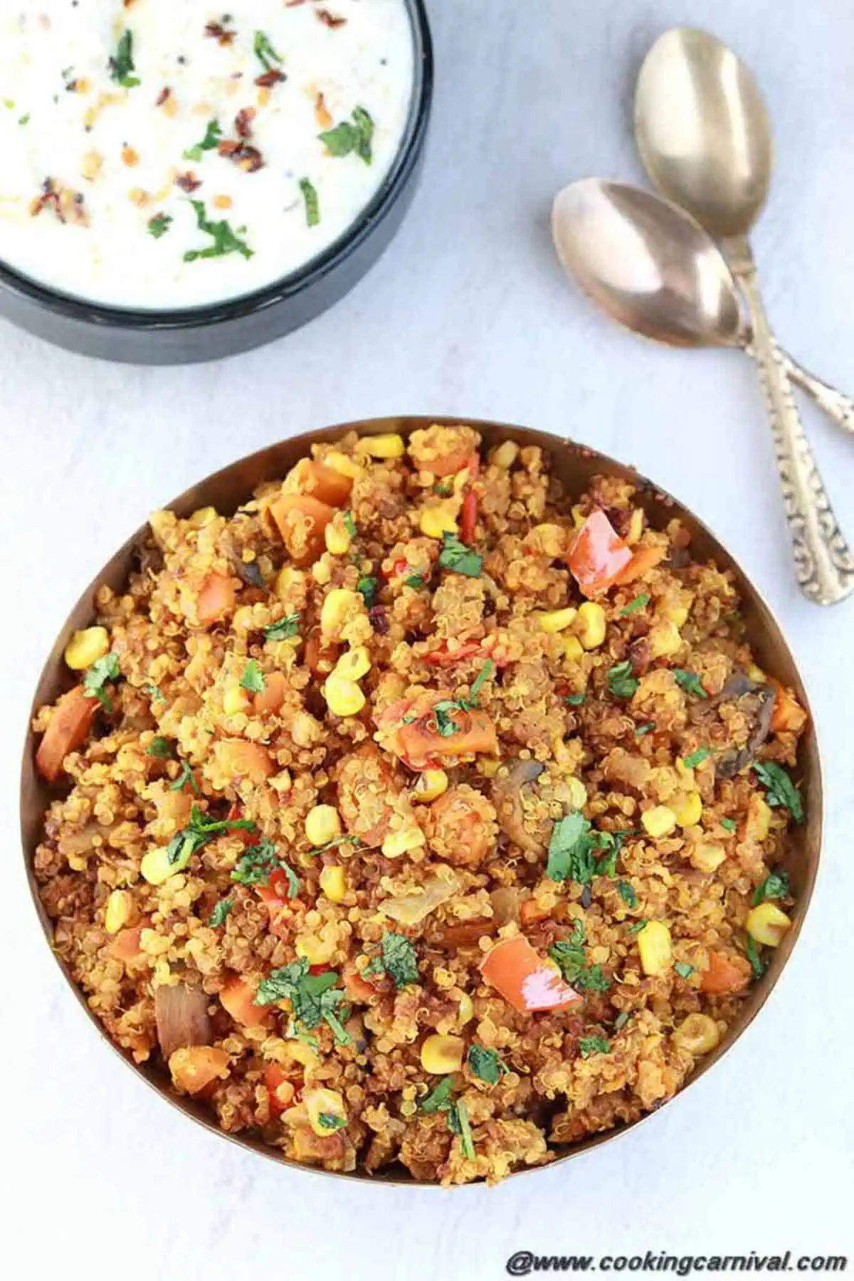 A Kansa bowl filled with vegetable quinoa pulav, spoons and raita on the side.