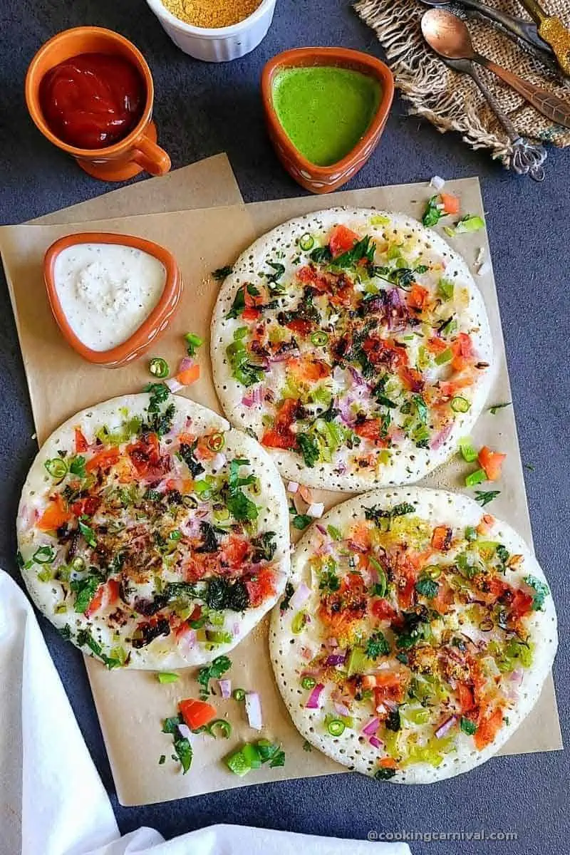 Masala Uttapam with coconut chutney, green chutney, tomato ketchup and some spoons, on gray board.